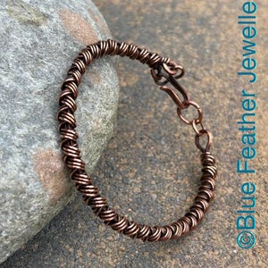 May include: A copper wire bracelet featuring a continuous coiled design with a hook and loop closure. The bracelet is metallic brown in color and displays the text Blue Feather Jewellery vertically along the right side of the image.