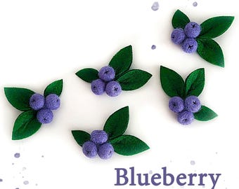 May include: Five felt blueberry clusters with green leaves on a white background with purple watercolour splatters. The word "Blueberry" is written in a dark purple font below the berries.