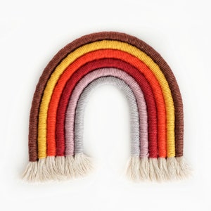 May include: A rainbow-coloured macrame wall hanging with a brown, yellow, orange, red, purple, and grey colour scheme. The macrame is made of thick yarn and has a fringe at the bottom.