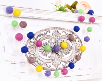Puede incluir: Una guirnalda de bolas de fieltro de colores con bolas moradas, azules, amarillas, verdes y grises enhebradas en una cuerda blanca. La guirnalda está colgada en una repisa blanca con un diseño metálico decorativo.