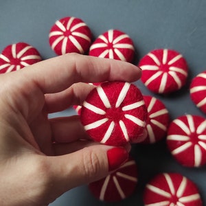 May include: Red felt balls with white stripes, resembling sweets, are scattered on a blue-grey surface. One is held in a hand. These handmade decorations could be used for crafts or festive displays, adding a touch of colour.