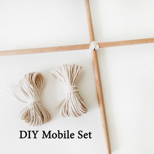 Puede incluir: Un kit de móvil de bricolaje con dos carretes de cuerda natural y dos listones de madera. Los listones están cruzados en el centro y atados juntos con cuerda. El texto "DIY Mobile Set" está impreso debajo de la imagen.