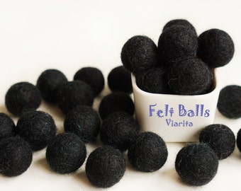 May include: A white bowl filled with black felt balls, with the words "Felt Balls Viarita" printed on the front of the bowl. There are more black felt balls scattered around the bowl on a white surface.