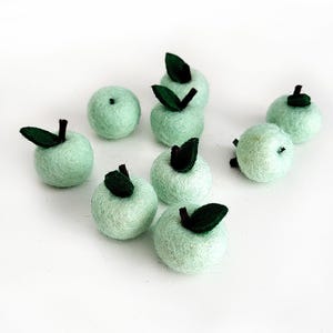Pode incluir: Uma coleção de maçãs de feltro feitas à mão em uma cor verde suave. Cada maçã tem uma folha de feltro verde escuro e um pequeno caule marrom. As maçãs estão espalhadas em uma superfície branca.