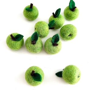 Pode incluir: Uma coleção de maçãs verdes de feltro feitas à mão com caules castanhos e folhas verdes de feltro. As maçãs estão espalhadas sobre um fundo branco, mostrando sua cor e textura vibrantes. Ideais para projetos de artesanato.