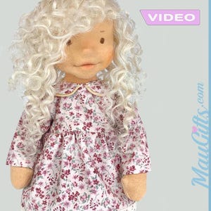 May include: A fabric doll with a detailed face and long, curly, white hair. The doll is dressed in a floral print dress with a Peter Pan collar. The dress has a pink and red floral pattern on a white background. The doll's body is soft.