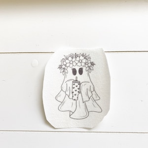 May include: A black and white illustration of a ghost wearing a flower crown and holding a cup with a straw. The ghost has two large eyes and a small mouth.