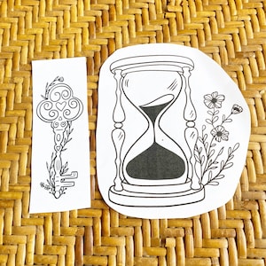 May include: Two black and white line art illustrations on white paper. One is a key with floral accents, the other is an hourglass with black sand and floral accents. The images are on a woven, light brown surface.