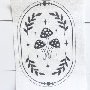 May include: Black and white illustration of three mushrooms with white spots inside a circular frame with a leafy border and moon and star accents.