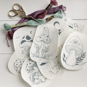 May include: A set of eight mushroom embroidery patterns on white fabric. The patterns feature various mushroom designs, including a mushroom with an eye, a mushroom with polka dots, and a mushroom with a crescent moon. The patterns are ready for embroidery with floss.