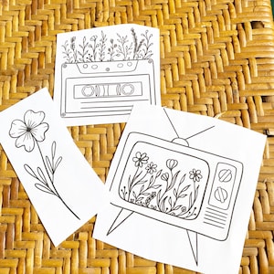 May include: Three black and white line drawings on white paper. The first drawing is a cassette tape with flowers growing out of it. The second drawing is a single flower with a stem. The third drawing is a television with flowers growing inside of it.