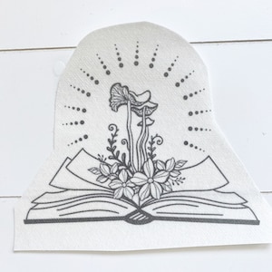 May include: A monochrome illustration featuring an open book with floral and mushroom elements. A dotted sunburst design frames the image, offering a whimsical, nature-themed aesthetic. The artwork is detailed and intricate.
