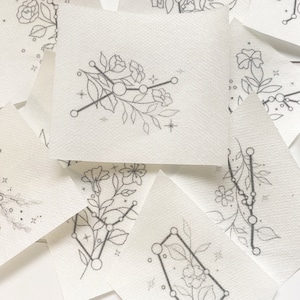 May include: Several small, square, white paper pieces with fine black line drawings. Each piece displays floral and constellation designs, including flowers, leaves, and star patterns. The overall aesthetic is minimalist and artistic.