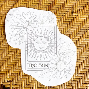 May include: A black and white illustration of a tarot card depicting the sun. The card features a stylised sun with a face and rays, surrounded by flowers. The text "The Sun" is printed at the bottom of the card.
