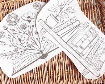 May include: Two white fabric patches with gray line drawings. One shows an open book with flowers and foliage. The other depicts a bookshelf with books and a potted plant. The patches are set on a woven wicker surface.