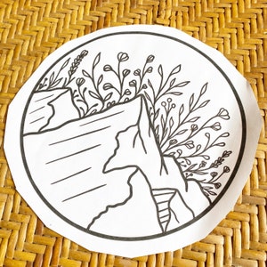 May include: Black and white line drawing of a mountain range with a cluster of flowers and leaves in the foreground. The drawing is on a white circular background.