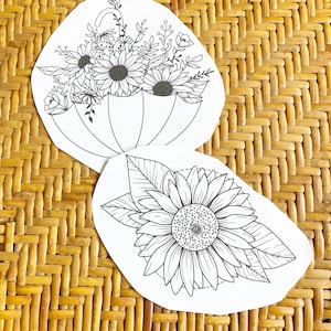 May include: Two black and white line drawings of flowers on white paper. The first drawing is of a bouquet of flowers under an umbrella. The second drawing is of a sunflower.