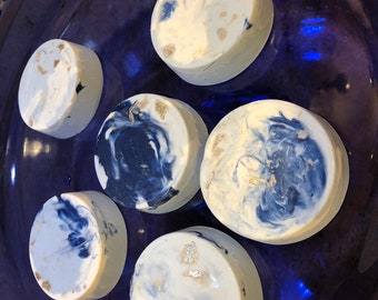 May include: Six round, handmade soaps with a marbled design in white, navy blue, and gold flecks. The soaps are arranged on a dark blue, decorative glass plate. The soaps have a smooth, rounded shape.