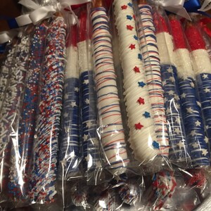May include: A collection of pretzel rods dipped in white chocolate and decorated with red, white, and blue sprinkles and stars. The pretzels are individually wrapped in clear plastic bags and tied with white ribbon.