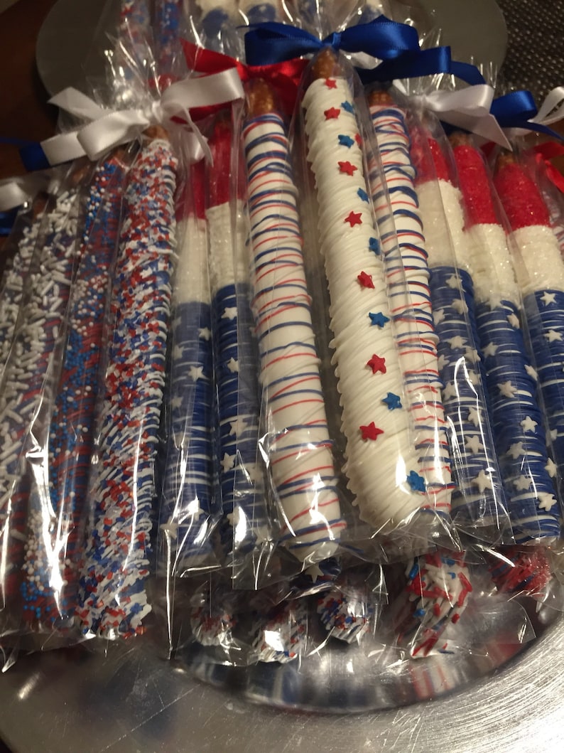 May include: A collection of pretzel rods dipped in white chocolate and decorated with red, white, and blue sprinkles and stars. The pretzels are individually wrapped in clear plastic bags and tied with white ribbon.