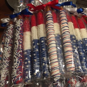 May include: A group of pretzel rods dipped in white chocolate and decorated with red, white, and blue sprinkles and designs. The pretzels are individually wrapped in clear plastic bags and tied with red, white, and blue ribbons.