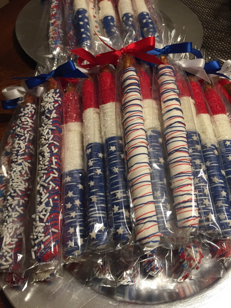 May include: A group of pretzel rods dipped in white chocolate and decorated with red, white, and blue sprinkles and designs. The pretzels are individually wrapped in clear plastic bags and tied with red, white, and blue ribbons.