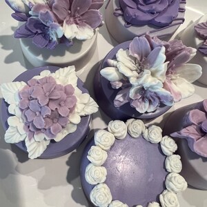 May include: Six purple and white decorated cookies with various floral designs. The cookies are arranged on a white surface.