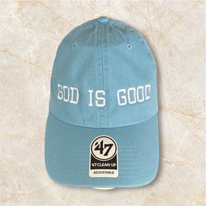 May include: A light blue baseball cap with white embroidered text that reads "God is Good". The cap has a black and white 47 Clean Up adjustable label.
