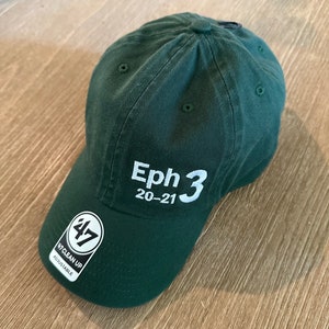 May include: A dark green baseball cap with a white embroidered logo that says "Eph 3 20-21". The cap has a black adjustable strap with a white "47" logo and the words "Clean Up Adjustable" printed on it.