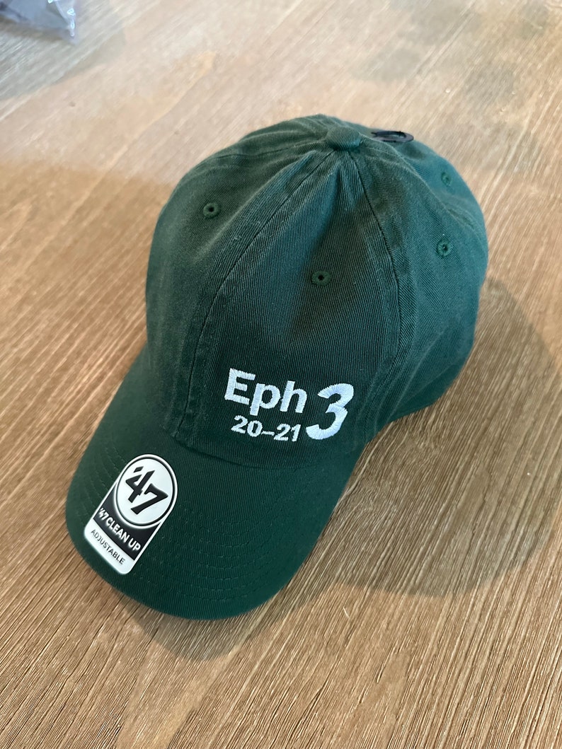 May include: A dark green baseball cap with a white embroidered logo that says "Eph 3 20-21". The cap has a black adjustable strap with a white "47" logo and the words "Clean Up Adjustable" printed on it.