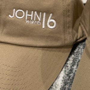 May include: A tan baseball cap with white embroidered text that reads "JOHN 16 THREE".