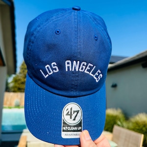 May include: A blue baseball cap with white embroidery that reads "Los Angeles" and a white "47 Clean Up Adjustable" logo on the side.