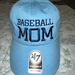 May include: A light blue baseball cap with the text "BASEBALL MOM" embroidered in navy blue. The cap has a white label with the text "47 CLEAN UP ADJUSTABLE" and the number "47" in a circle.