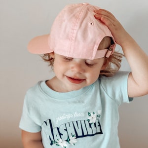 May include: A young child wearing a light blue t-shirt with the text "greetings from Nashville" and white flowers. The child is wearing a pink baseball cap.