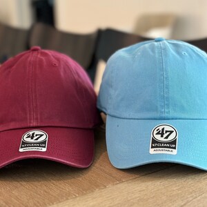 May include: Two baseball caps, one maroon and one light blue, both with a white logo that says "47 Clean Up Adjustable".