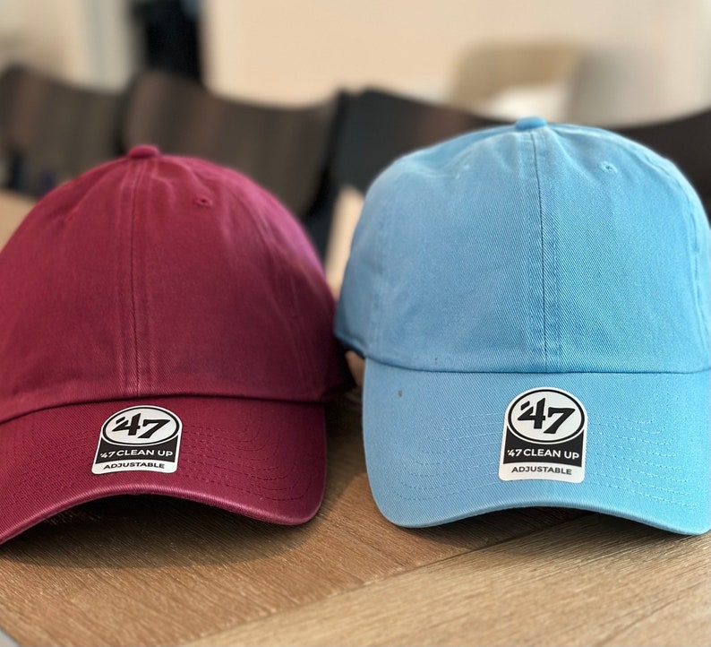 May include: Two baseball caps, one maroon and one light blue, both with a white logo that says "47 Clean Up Adjustable".