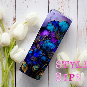 May include: A stainless steel tumbler with a floral design in shades of blue, purple, and gold. The tumbler is decorated with a watercolor-style floral pattern and the text "STYLISH SIPS SK". White tulips are arranged around the tumbler.