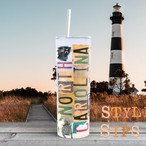 May include: A stainless steel tumbler with a straw, featuring the text "NORTH CAROLINA" in a collage of license plates and a Carolina Panthers logo. The background includes a lighthouse and a wooden walkway.