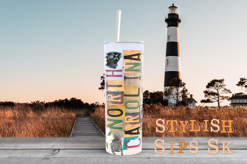 May include: A stainless steel tumbler with a straw, featuring the text "NORTH CAROLINA" in a collage of license plates and a Carolina Panthers logo. The background includes a lighthouse and a wooden walkway.