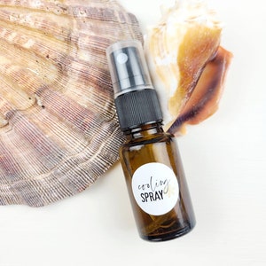 May include: A small brown glass bottle with a black spray top and a white round label that says "cooling spray" in black script. The bottle is sitting on a white surface next to a large seashell.