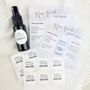 May include: A white background with a black and white label that reads "Warm Vanilla Room Spray". The label is on a small, clear glass bottle with a black spray nozzle. There are also several white round labels with the same text on them.