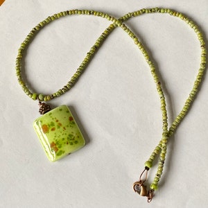 May include: A green and brown speckled ceramic pendant hangs from a green beaded necklace. The necklace has a bronze clasp.