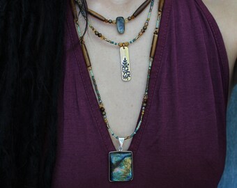 Puede incluir: Un collar de capas con múltiples hileras de cuentas y colgantes. El collar presenta un colgante cuadrado con una piedra iridiscente azul y verde, un colgante rectangular con un diseño de árbol y cuentas de madera marrón.