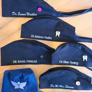 May include: Six black scrub caps with different names embroidered on them. The names are Dr. Emma Winkler, Dr. Isidoro Ferlito, DR. RAHUL IYENGAR, Dr. Ellen Tsang, and Dr. W. Alex Stinson. Each cap has a different button or pin on it.
