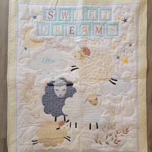 May include: A quilted baby blanket with a cream-colored background and a light yellow border. The blanket features embroidered sheep, clouds, stars, and the words "SWEET DREAMS" in pastel colors. The name "Ellie" is also embroidered on the blanket.
