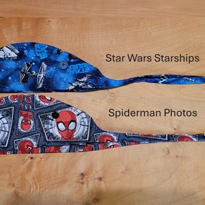 Könnte beinhalten: Zwei OP-Hauben. Eine ist blau mit einem Star Wars Starships-Design. Die andere ist grau mit einem Spider-Man Photos-Design. Beide Kappen haben einen Knopf oben und einen langen Schwanz.