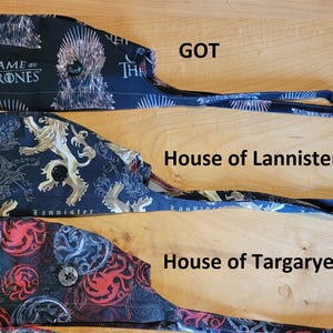 May include: Three black scrub hats with different Game of Thrones designs. The top hat features the Iron Throne and the text "Game of Thrones". The middle hat features a gold lion and the text "Lannister". The bottom hat features a red dragon and the text "House Targaryen".