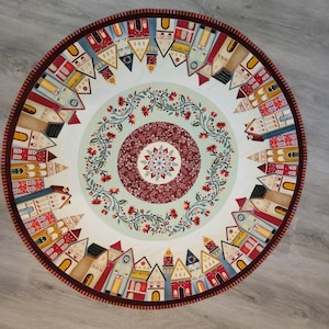 May include: A circular fabric mat featuring a central medallion with a red floral pattern, surrounded by a light green ring with a red floral vine border. The outer edge displays a colorful array of stylized houses in red, yellow, and blue.