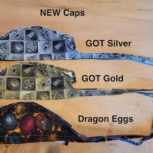 May include: Four different patterned fabric scrub caps with ties. The caps are labeled "GOT Silver", "GOT Gold", "Dragon Eggs", and "NEW Caps".