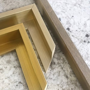 May include: Two picture frames, one with a gold finish and one with a light brown finish. The frames are made of wood and have a simple, classic design.